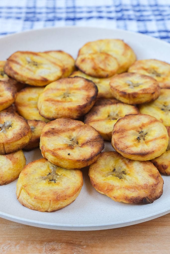 crispy plantain slices on white plate with blue cloth behind on wooden board.