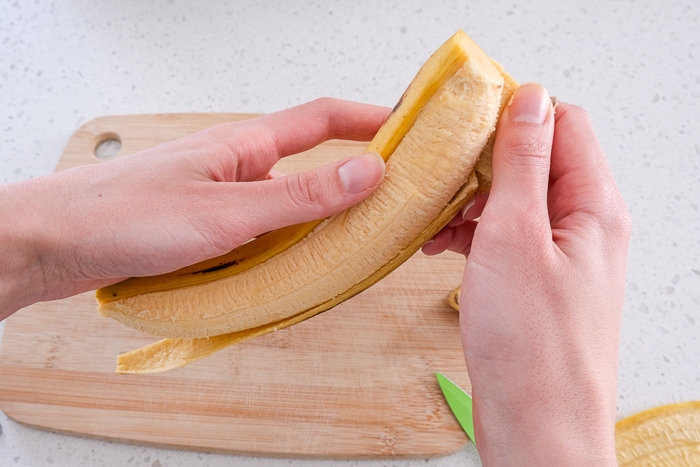 hands peeling peel off of yellow plantain above wooden cutting board.