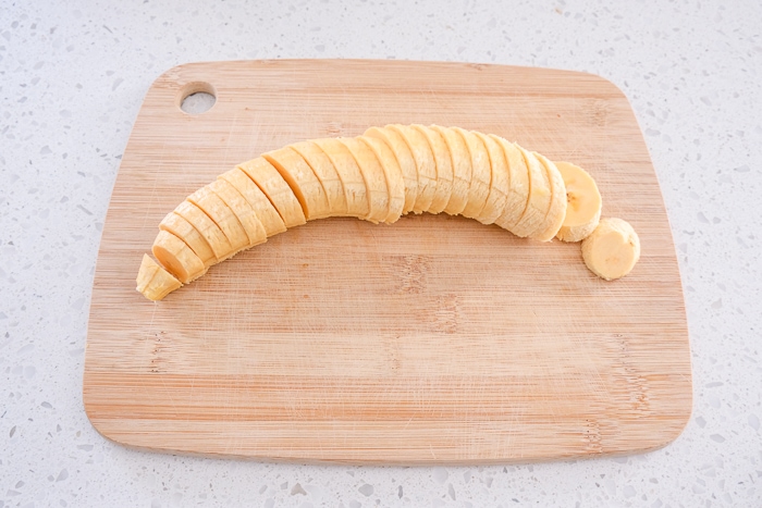 unpeeled plantain cut into slices on wooden board on white counter.