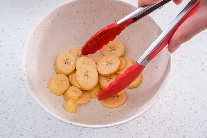 red tongs mixing plantain slices in white bowl on counter.