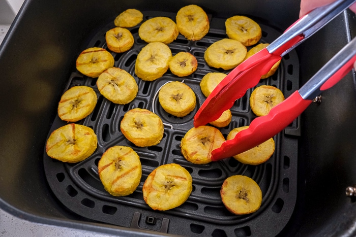 red tongs flipping crispy plantain chips in black air fryer tray.