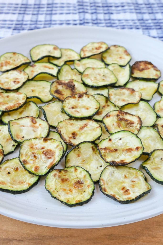 green zucchini chips on white plate with wood under and cloth behind.