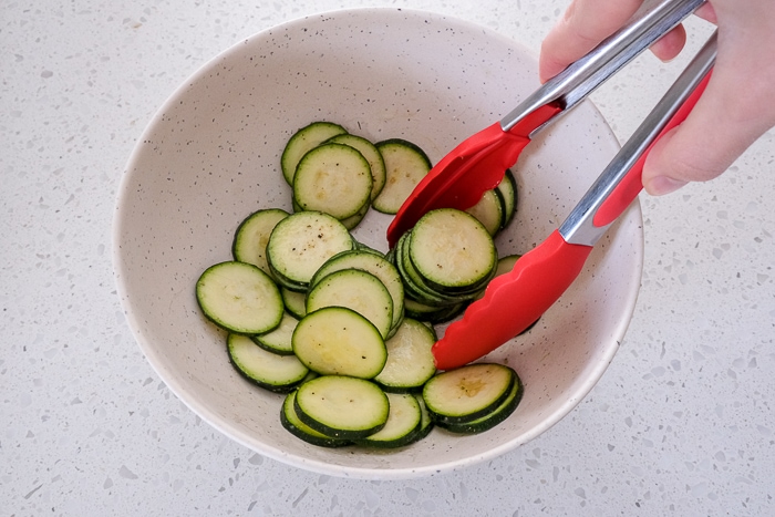red tongs mixing slices of zucchini in white bowl on counter top.