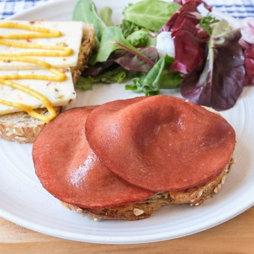 pieces of fried bologna on bread with cheese and salad behind all on white plate.