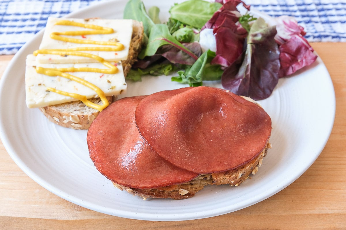pieces of fried bologna on bread with cheese and salad behind all on white plate.