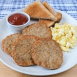 cooked sausage patties on white plate with eggs and toast behind.