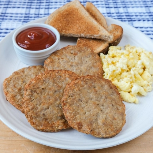 cooked sausage patties on white plate with eggs and toast behind.