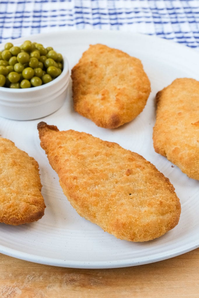 breaded fish fillets on white plate with bowl of peas beside on wooden board with cloth behind.