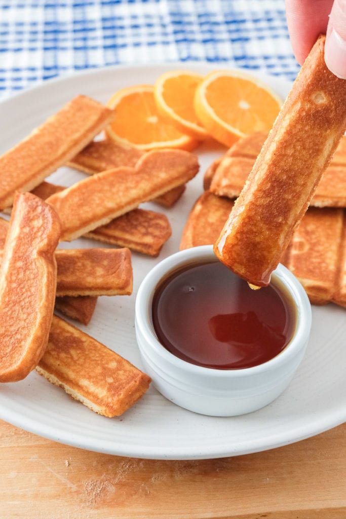 hand dipping french toast stick into white cup of syrup with french toast sticks and orange slices behind on plate.