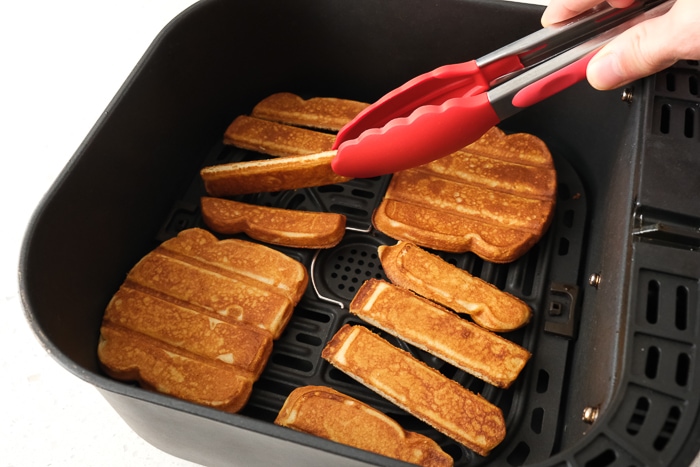 red tongs flipping french toast sticks in air fryer tray on counter top.