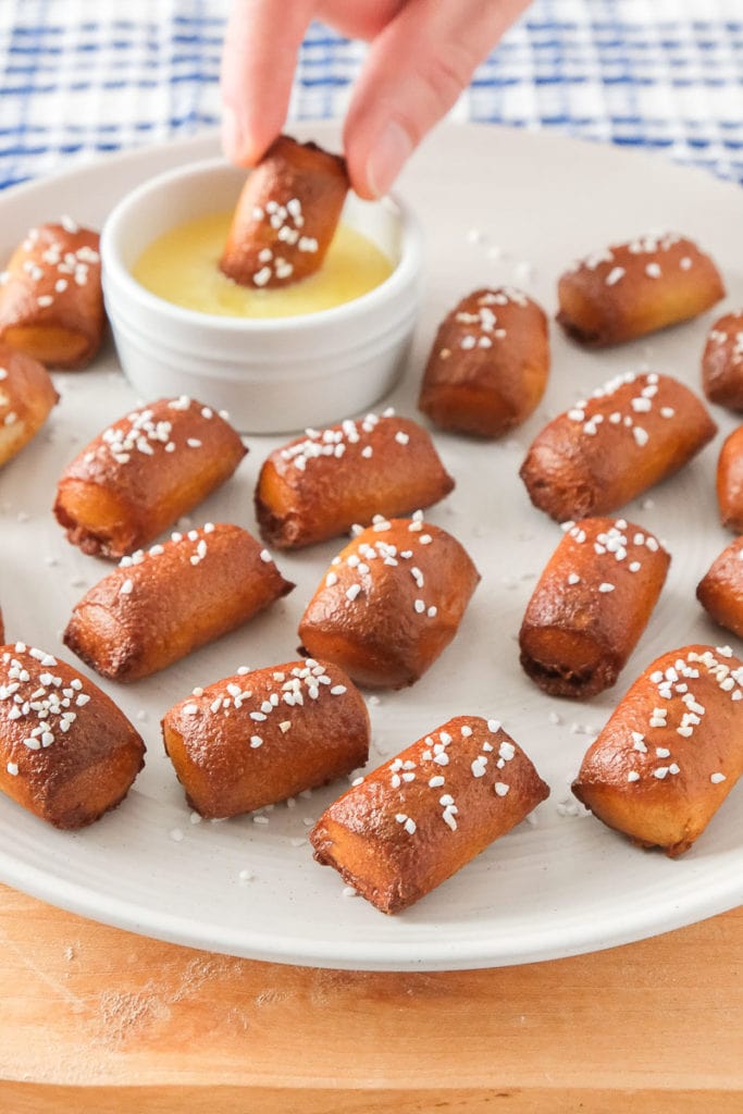 hand dipping pretzel bite into butter behind white plate of many pretzel bites on wooden board.