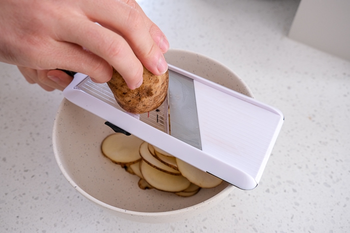 hand holding potato over white mandolin slicer with potato slices in bowl underneath.