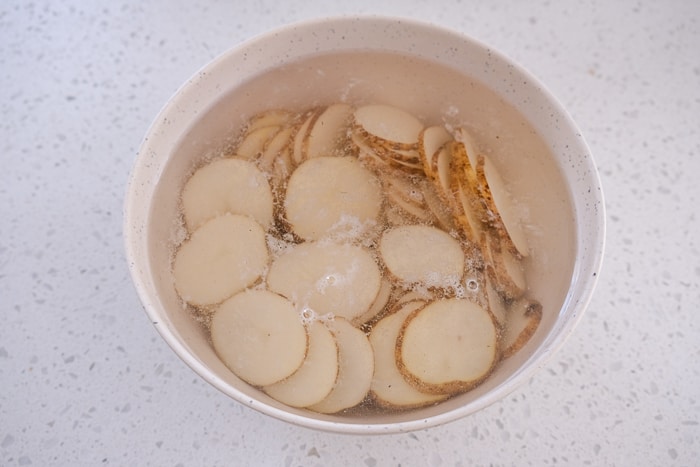 potato slices in white bowl full of water on white counter top.