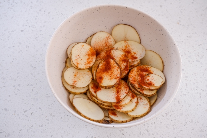 red spices on raw potato slices in white bowl on white counter.