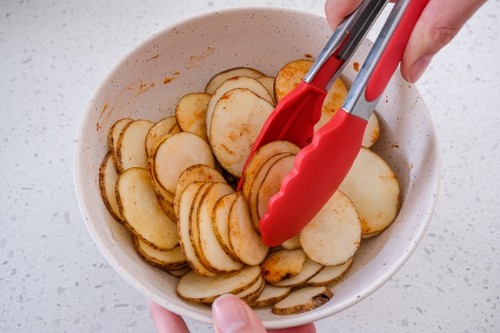 red tongs mixing potato slices in white bowl on white counter.