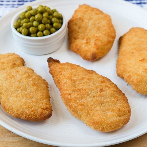 crispy breaded fish on white plate with green peas beside all on wooden board.