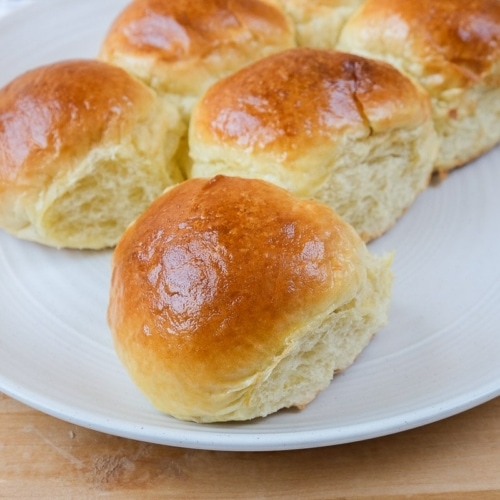 cooked dinner rolls on white plate with wooden board under and cloth behind.