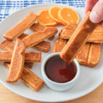 hand dipping french toast stick into syrup on white plate with orange slices behind.