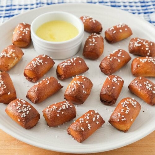 crispy pretzel bites covered in salt on white plate with butter behind on wooden board.
