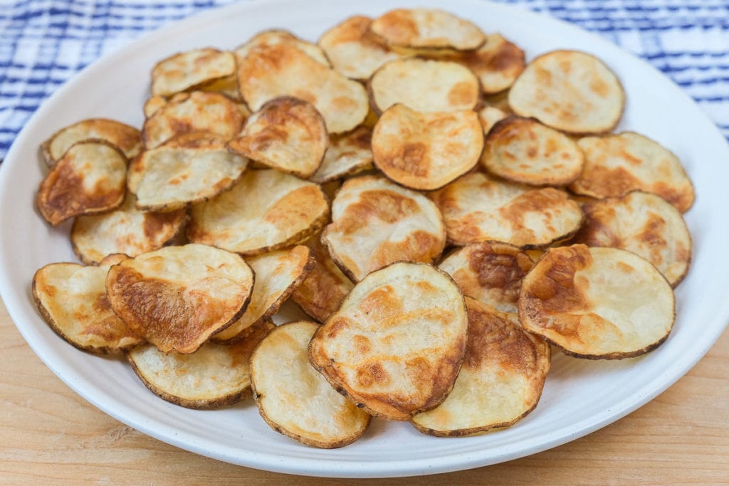 crispy potato chips on white plate with wood under and cloth behind.