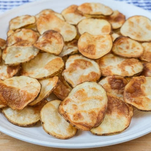 crispy potato chips on white plate with wood under and cloth behind.