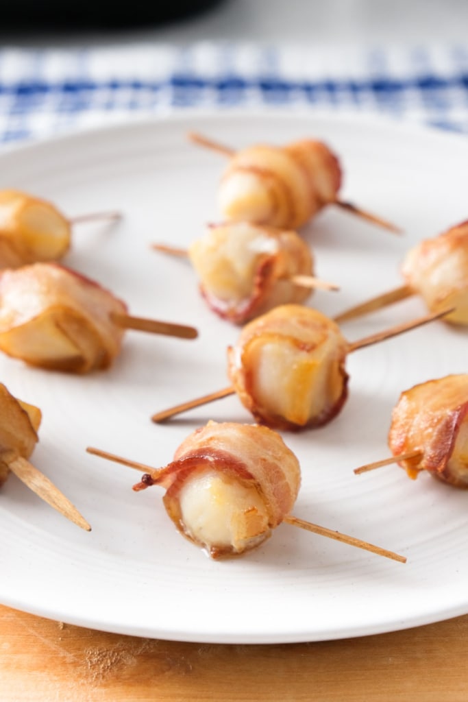 scallops wrapped in bacon with a wooden skewer through them on on white plate sitting on wooden board.