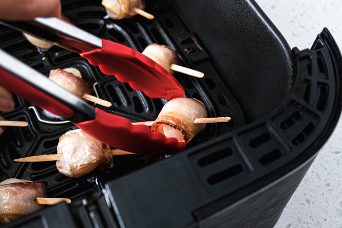 red tipped tongs turning scallop wrapped in bacon above air fryer tray on counter.