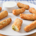 crispy breaded pickles on white plate with bowl of white dip behind.
