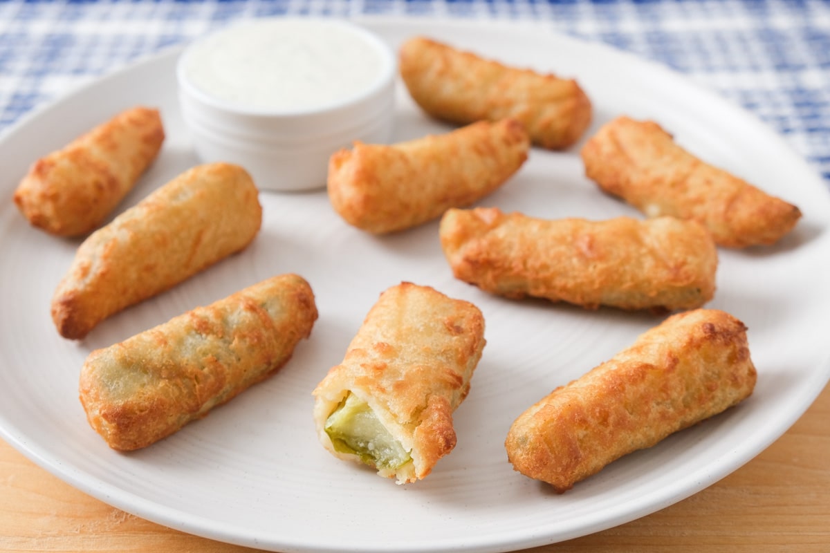 crispy breaded pickles on white plate with bowl of white dip behind.