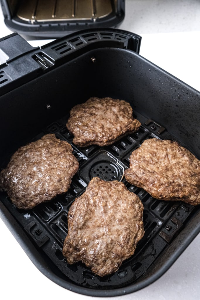 four cooked hamburger patties in black air fryer tray on white counter with air fryer behind.
