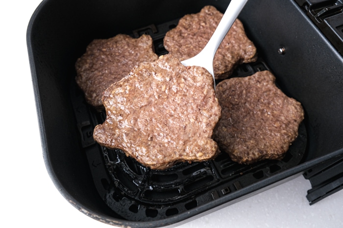 white flipper under cooked burger patty above other cooked patties sitting in black air fryer tray.