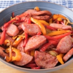 blue bowl of cooked kielbasa and peppers on wooden board with blue towel behind.
