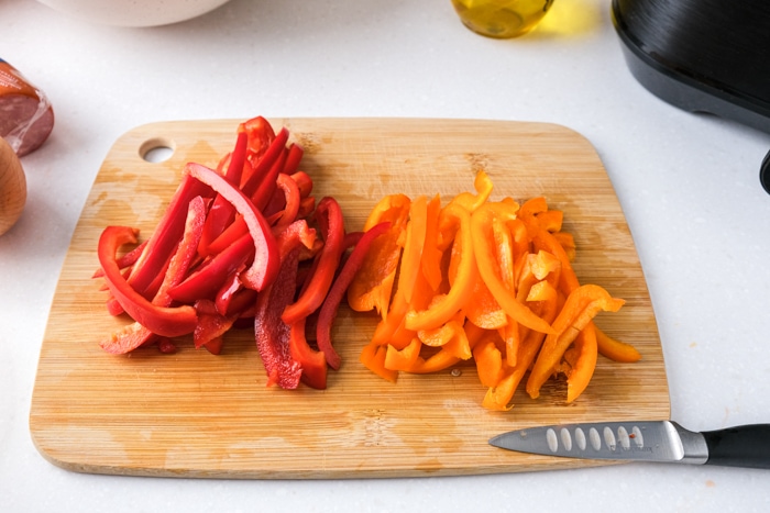 strips of red and orange peppers cut on wooden board with knife in front on white counter.