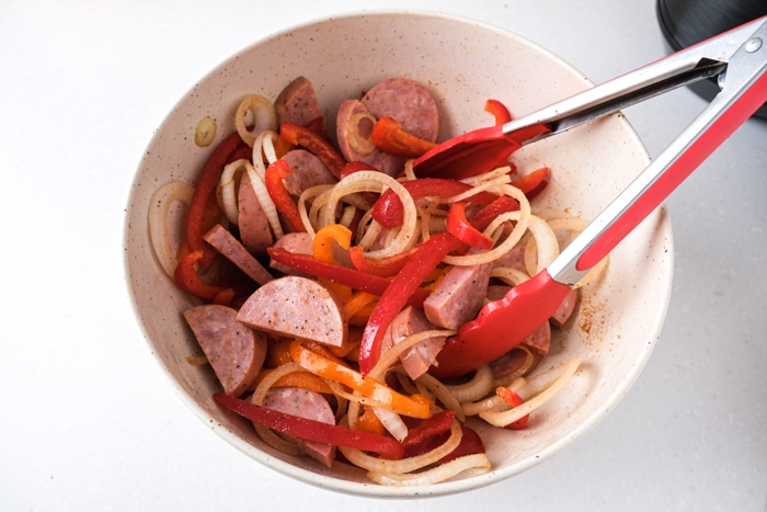 kielbasa onions and peppers in white bowl bring mixed with red tipped tongs.