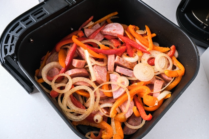uncooked peppers onions and chunks of kielbasa sitting in black air fryer tray on white counter.