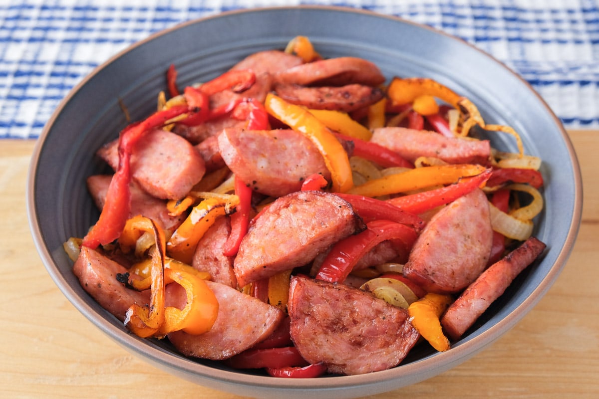 crispy kielbasa with red and yellow pepper pieces in blue serving bowl sitting on wooden cutting board.