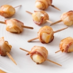 crispy bacon wrapped scallops with wooden sticks through on white plate.