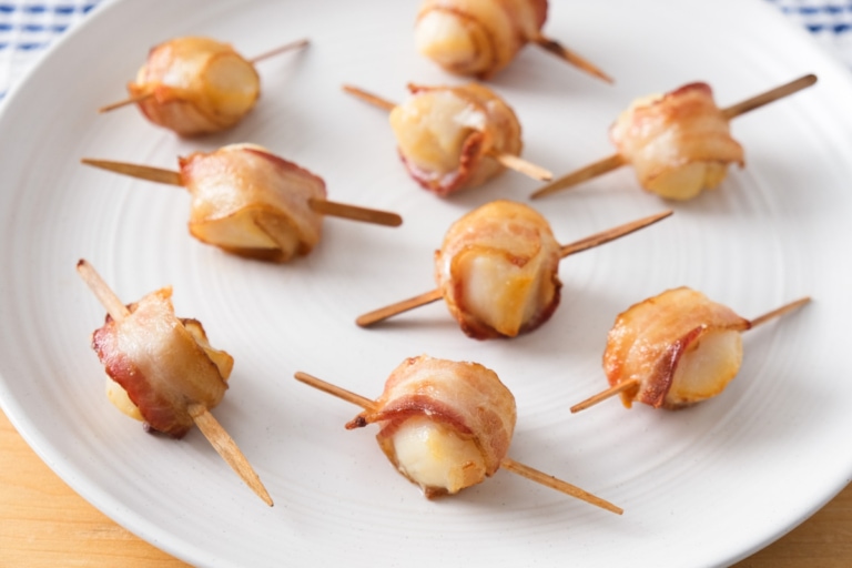 crispy bacon wrapped scallops with wooden sticks through on white plate.