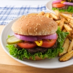 well dressed hamburger with sesame seed bun on plate with fries sitting on wooden board.