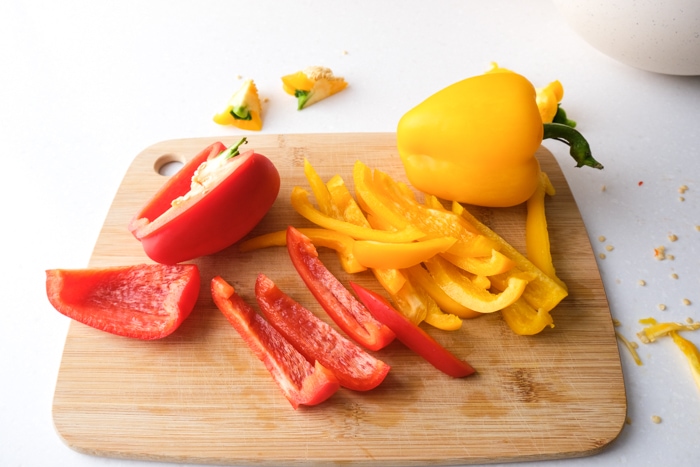 red and yellow bell peppers cut into strips sitting on wooden cutting board on white counter.