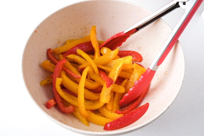 red tipped tongs mixing raw bell pepper slices and spices in white mixing bowl on counter.