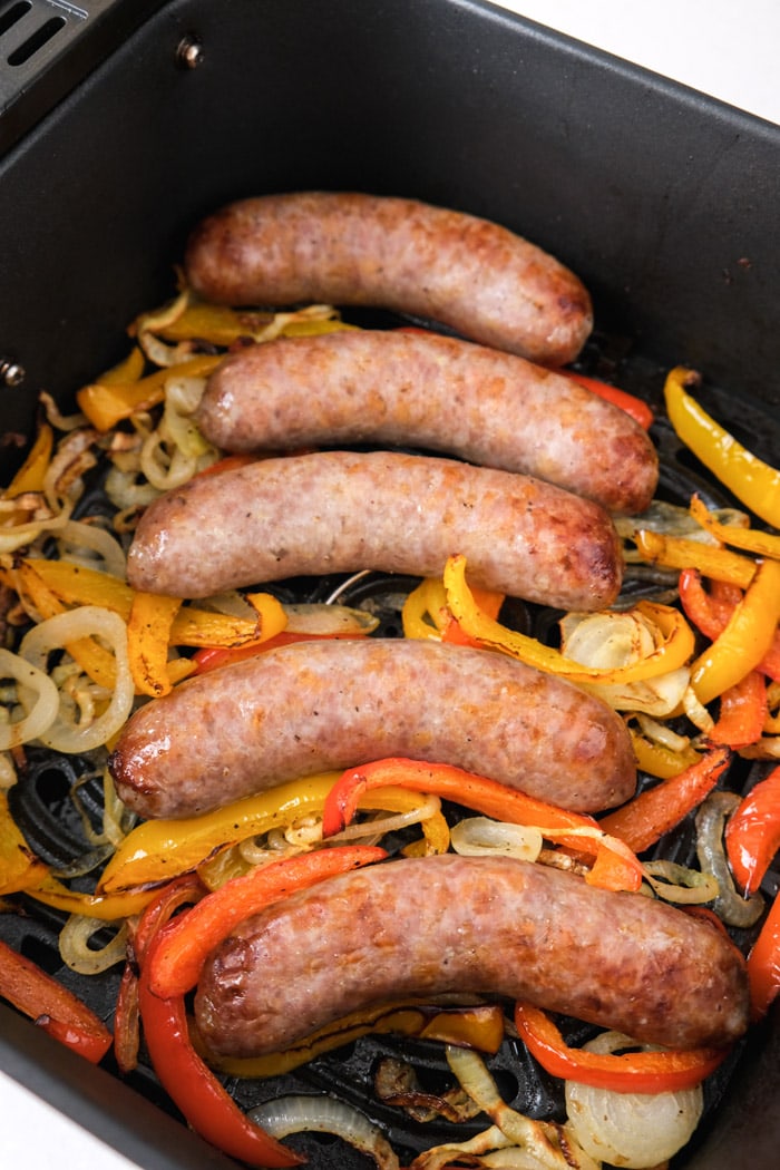 cooked brat sausages on bed of cooked peppers in black air fryer tray on counter.