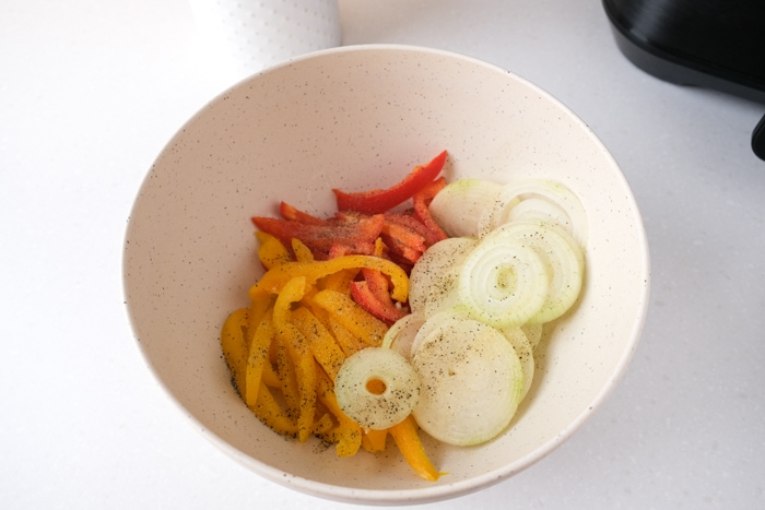 slices of white onion and strips of bell peppers covered in spices in white mixing bowl on counter.