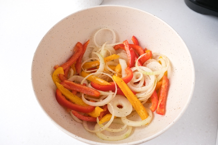 bell pepper strips and onions coated in oil and spices in white bowl on white counter top.