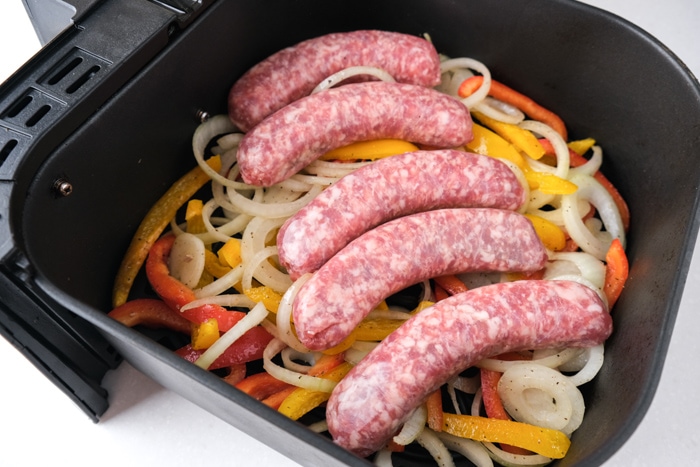 raw brat sausages on bed of raw onions and peppers in black air fryer basket.