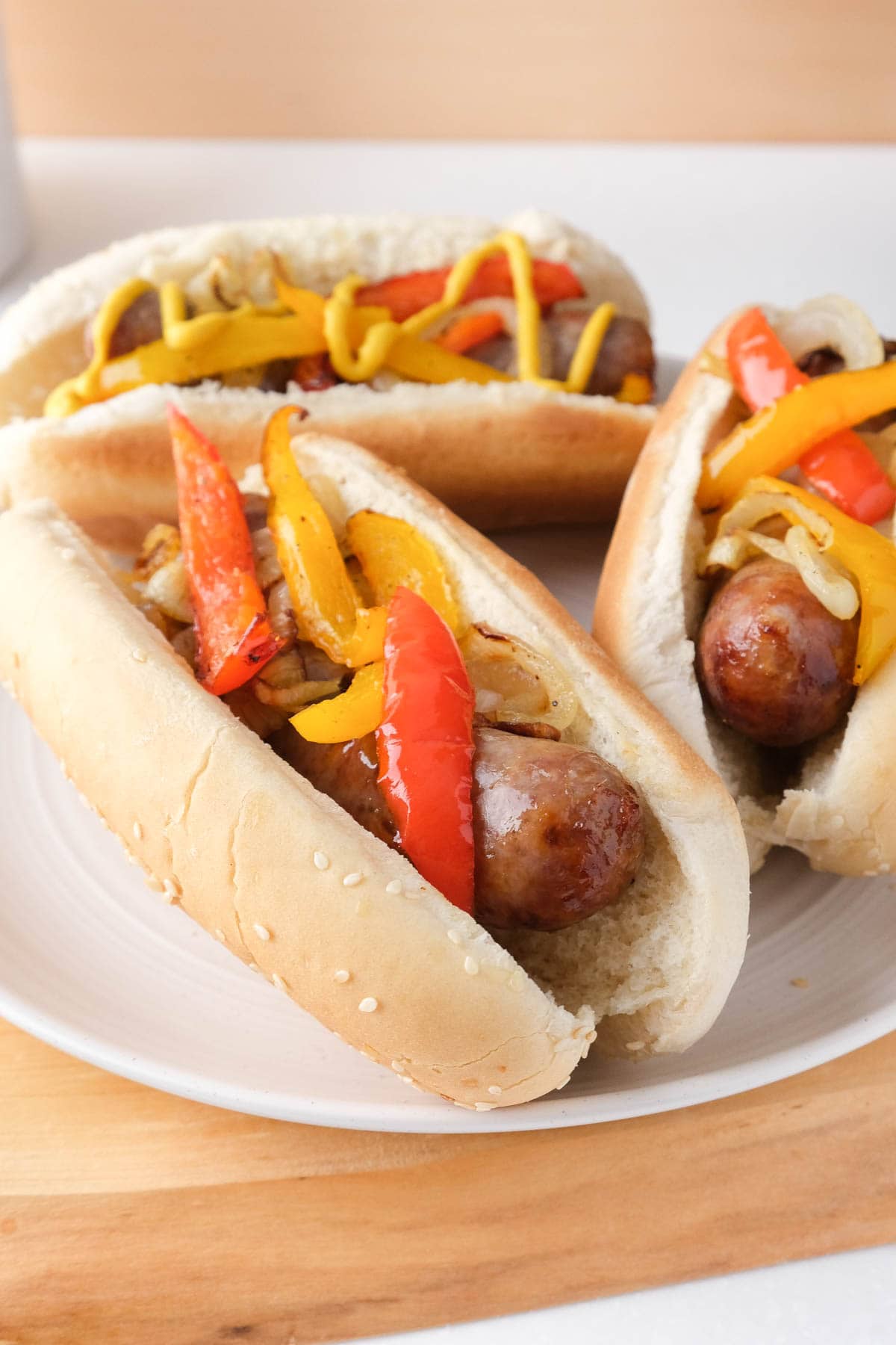 brats in buns with grilled peppers on top on white plate with wooden board under.