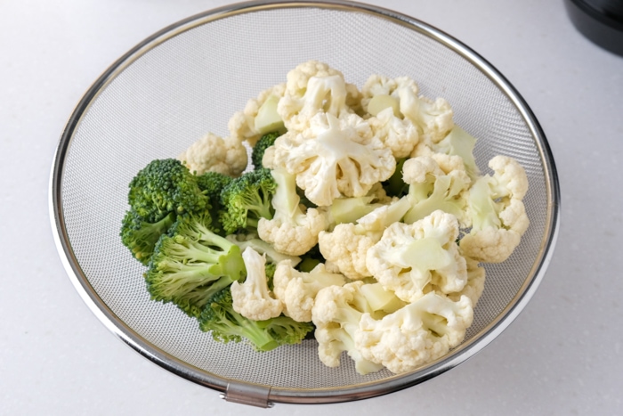 small cut broccoli and cauliflower florets in metal strainer on white counter.