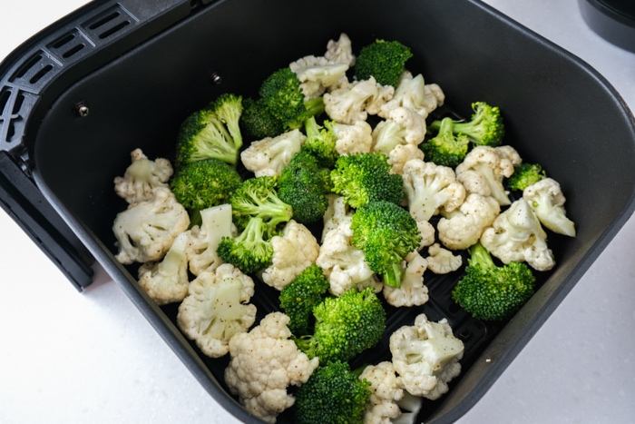 raw broccoli and cauliflower florets in black air fryer basket on white counter.