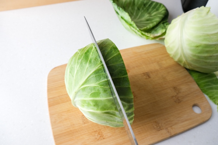 long knife cutting large slice off head of cabbage all on wooden cutting board.