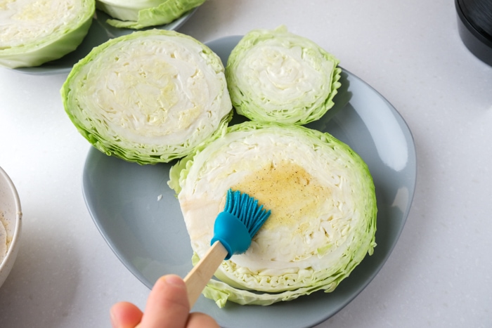 blue tipped cooking brush brushing oil and spices on raw cabbage steaks on blue grey plate on counter.